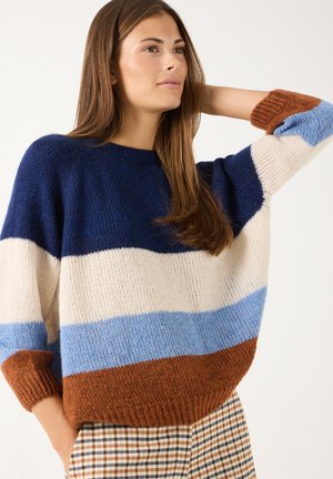 Vrouw met lang bruin haar die een gebreide trui draagt met brede horizontale strepen in marineblauw, wit, lichtblauw en bruin, poserend met één hand achter haar hoofd.