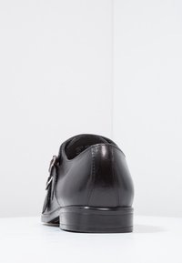 Chaussure de costume pour homme en cuir noir avec double boucle vue de dos sur une surface blanche contre un fond blanc uni.