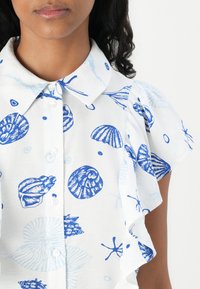 Hvit bluse med blå skjell og hav-tema mønstre. Knappefront med krage og volangerm. Mykt, lett stoff.