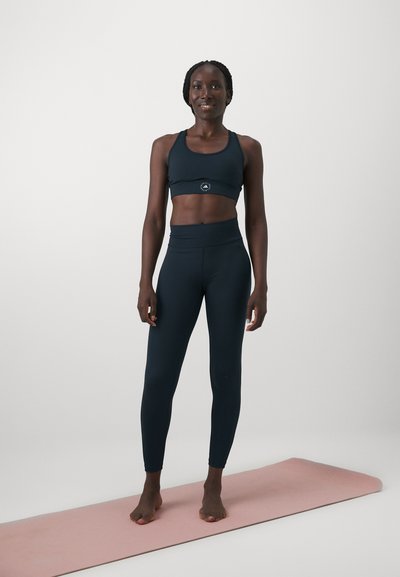 Ναυτικό σουτιέν γυμναστικής και ψηλόμεσα κολάν από ελαστικό ύφασμα, φορεμένα από ένα άτομο που στέκεται σε ροζ στρώμα γιόγκα.