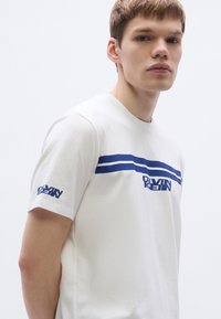 Bela bombažna majica z modrimi vodoravnimi črtami ter logotipom na prsih in rokavu. Okrogel izrez in kratki rokavi. Gladek otip.