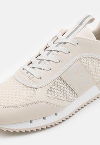 Beige sneaker with mesh details, white laces, a grey stripe on the tongue, and "EA7 Emporio Armani" logo on the side.