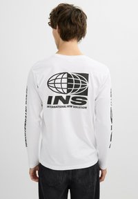 Camiseta de manga larga blanca con logo de globo negro y texto "INS" en la parte posterior; acentos gráficos a lo largo de ambas mangas. Material de algodón suave.