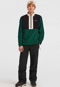 Pullover in pile verde, nero e beige con collo a mezza zip, indossato con pantaloni neri e stivali marroni; presenta un logo sul petto.