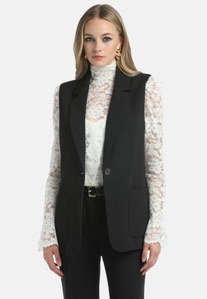 Gilet nero su misura con rever a scollo, due tasche frontali e chiusura con un solo bottone, abbinato a un top in pizzo bianco e pantaloni neri.