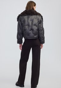 Kobieta w czarnej pikowanej kurtce z puszystym kołnierzem i mankietami, w połączeniu z czarnymi spodniami o szerokich nogawkach i ciemnym obuwiem, stojąca tyłem.