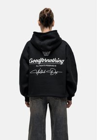Felpa nera oversize con una grande grafica bianca sul retro, che presenta un logo e una firma. Realizzata in tessuto spesso con cappuccio regolabile.