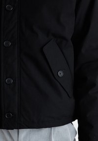 Black jacket with a textured finish, featuring six round buttons, a side pocket with flap, and contrasting stitching at the hem.
