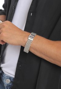 Silberne Metallarmbanduhr mit einem Gittermusterarmband und eckigem Zifferblatt, getragen an einem gebräunten Handgelenk, vor einem schwarzen Hemdhintergrund.