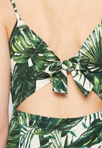 Top court vert et blanc avec un motif de feuilles tropicales, doté d'un lien au dos et de découpes triangulaires, fabriqué en tissu doux avec une texture lisse.