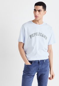 Svetlo modra kratka majica s kratkimi rokavi, na kateri je v sivi barvi natisnjen napis "PEPEJEANS" v loku. Kombinirana z temno modrimi kavbojkami, ki prikazujejo sproščen fit.