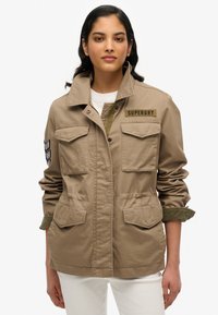 Lichtbruine militaire stijl jas van stof met vier voorzakken, een verstelbare taille en een "Superdry" logo op de borst.