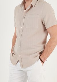 Camicia beige a maniche corte con bottone e tasca sul petto, realizzata in tessuto leggero, con un sottile motivo a trama e bottoni bianchi.