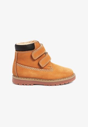 Sandy-farbene Wildleder-Ankle Boots mit zwei schwarzen Klettverschlüssen, einem mit Fell gefütterten Kragen und einer Gummisohle mit kleinen Profilmustern.