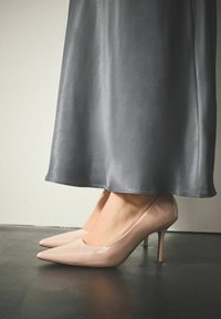 Naakte lakleren hoge hakken met een spitse neus en een dunne spijkerstilet, gecombineerd met een vloeiende grijze satijnen rok.