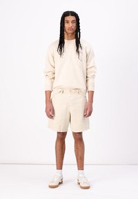 Light beige oversized sweatshirt with small red logo, paired with matching shorts. Model wears white sneakers and ankle socks. Plain background.