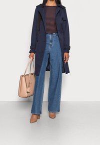 Cappotto trench navy sopra una maglietta a righe, jeans blu a gamba larga e scarpe beige con tacco. Borsa marrone con etichetta, texture morbida, design semplice.
