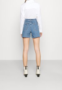 Pantalones cortos de mezclilla con dobladillo deshilachado, diseño de talle alto y dos bolsillos traseros, combinados con una camisa blanca con cuello y botines beige hasta el tobillo.