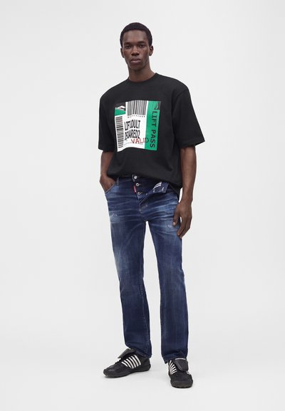 Άνδρας στέκεται φορώντας μαύρο oversized t-shirt με γραφικά, μπλε τζιν με κουμπιά και ετικέτα στη θηλιά της ζώνης, και μαύρα και λευκά αθλητικά παπούτσια σε λευκό φόντο.