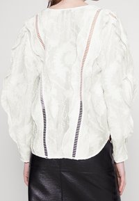 Blusa bianca con tessuto testurizzato, maniche a sbuffo e bordi arricciati. Presenta pannelli in pizzo verticali e un orlo arrotondato. Abbinata a una gonna nera.
