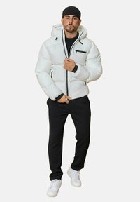 Doudoune blanche avec capuche, fermeture éclair noire et accents de poche, associée à un pantalon noir et des baskets blanches. Texture lisse, design souligné.