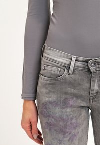 Camiseta gris de manga larga con un diseño ajustado, combinada con unos jeans gris desgastados que tienen un patrón floral en el muslo izquierdo y detalles de herrajes estándar.