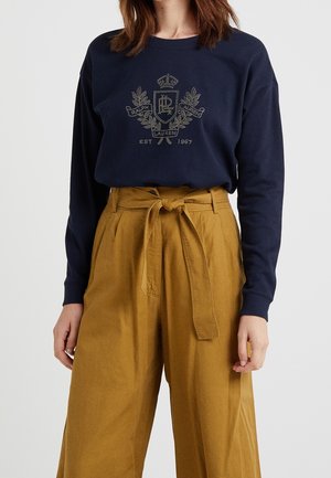 Personne portant un sweat-shirt bleu marine avec un motif de couronne de laurier et un pantalon large taille haute moutarde noué avec une ceinture en tissu.