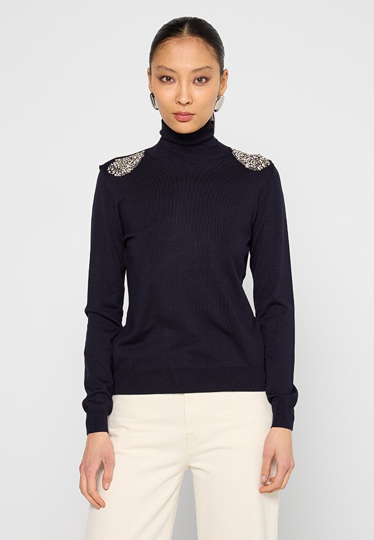 Maglione a collo alto blu navy con texture a coste, decorato con dettagli sui lati delle spalle e polsini aderenti. Abbinato a pantaloni chiari.