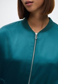 Teal selyem bomber dzseki poliészter felülettel, bordázott gallérral és ezüst cipzárral. Simább textúra és testhezálló dizájn.