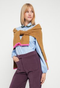 Brun strikket trøje med lyserøde detaljer lagt over en lyseblå skjorte; modellen har lilla højtaljede bukser med sidelommer.