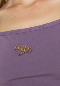 Lila tajt topp med en slät textur, med en upphöjd gul "Nike"-logotyp på bröstet. Framträdande med en bred halsringning.