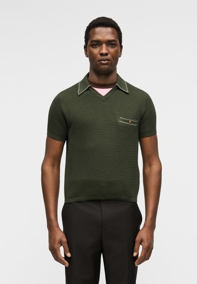 Άνδρας που φοράει ελαφρώς πράσινο πλεκτό πόλο με κοντά μανίκια και τσέπη στο στήθος, συνδυασμένο με μαύρο παντελόνι, στέκεται μπροστά σε απλό φόντο.