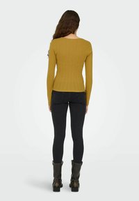 Sennepsgul ribbet langærmet top med en tætsiddende pasform, parret med mørke jeans og sorte ankelstøvler. Bagsiden har en scoop-udskæring.