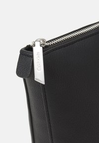 Pochette en cuir noir avec une finition texturée, dotée d'une fermeture éclair argentée et d'une languette, logo embossé et bords nets. Design compact.