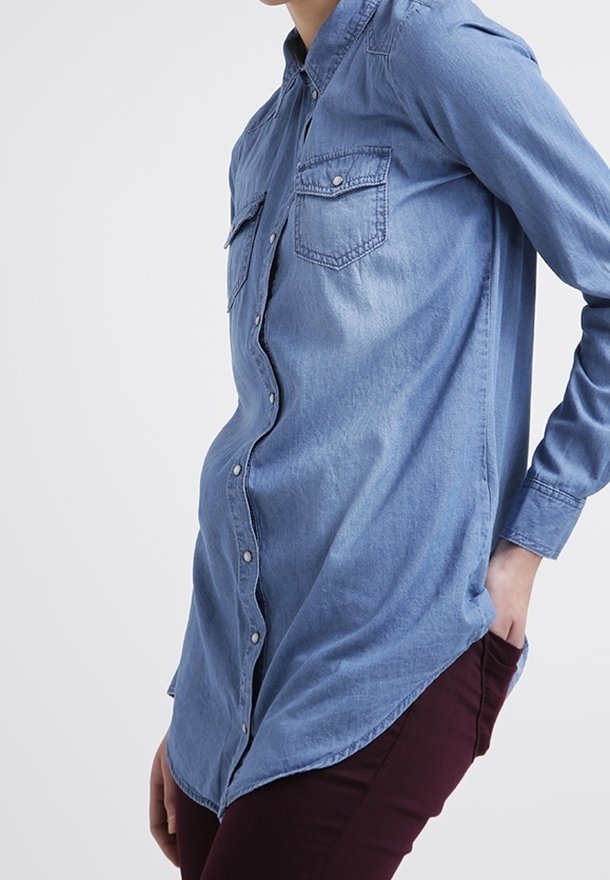 Ljusblå denimskjorta med långa ärmar, knäppning, och två bröstfickor. Har en rundad nederkant och en avslappnad passform.