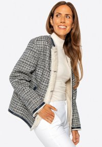 Tweed-Jacke mit einem strukturierten blau-weißen Muster, mit einem cremefarbenen Kunstpelz-Innenfutter und marineblauen Akzenten, getragen über einem hellen Pullover und weißen Hosen.