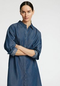 Tummansinisestä denimgarsista, jossa on napitus edessä, pitkät hihat, jotka on käännetty hihansuista, ja rento istuvuus. Sujuva pinta.