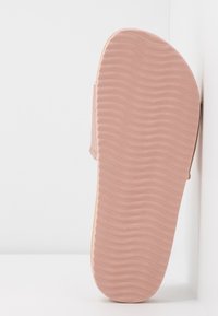 Semelle de sandale en caoutchouc rose unique avec un motif de bande de roulement ondulé, positionnée verticalement contre un mur et une surface blancs.