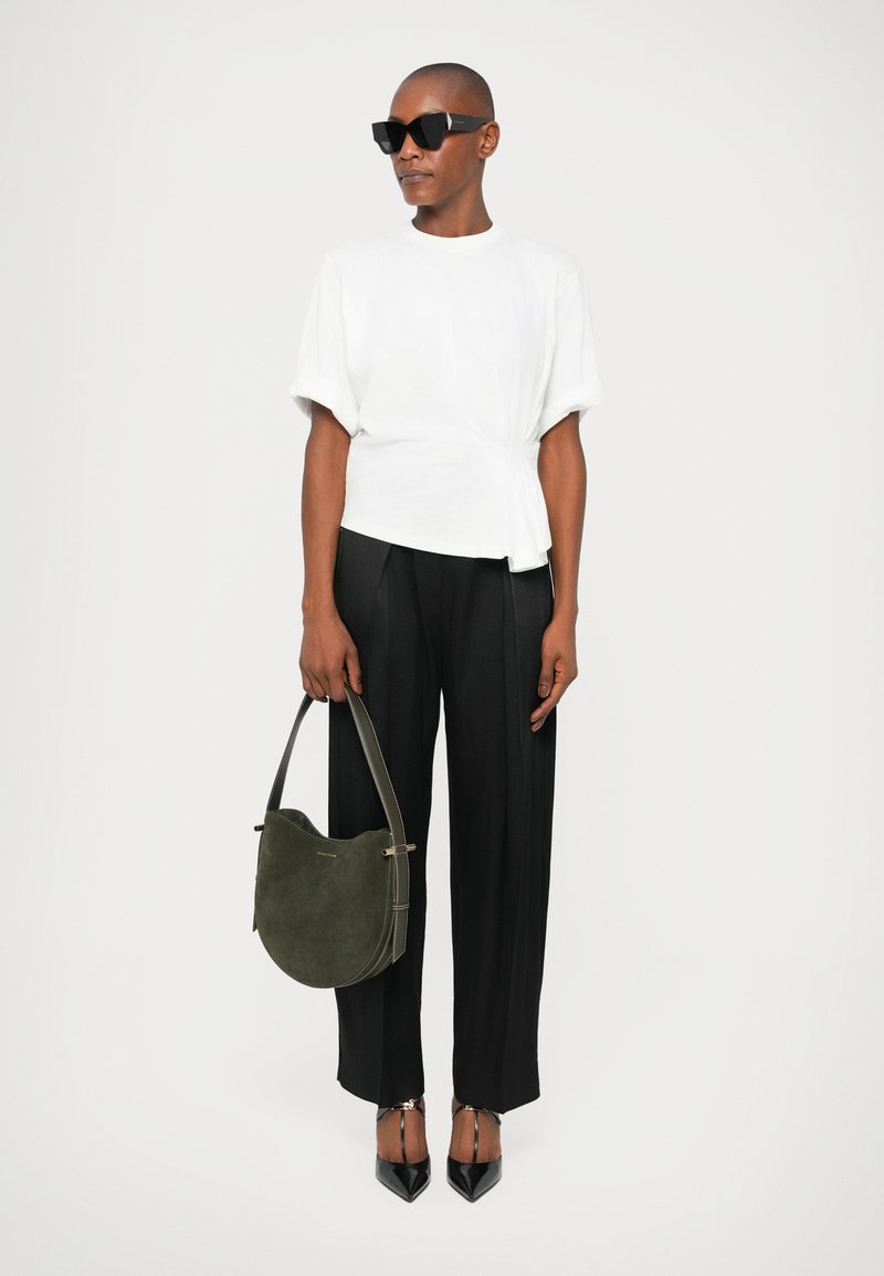 Top blanco de manga corta con detalle de nudo, pantalones negros de corte ajustado, bolso de ante verde, y tacones negros de punta, con gafas de sol.