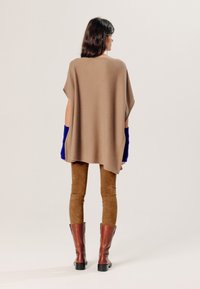 Beige poncho met korte mouwen, gecombineerd met bruine suède leggings en kniehoge bruine laarzen. De mouwen hebben blauwe accenten.