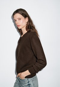 Brauner, gestrickter Cardigan mit Rundhalsausschnitt, vorderem Knopfverschluss und gerippten Bündchen. Kombiniert mit hellblauen Jeans.
