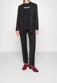 Blazer negro ajustado sobre una camiseta negra con el logo "BOSS", combinado con pantalones gris oscuro y mocasines burdeos.