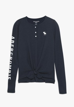 Chemise en maille gaufrée à manches longues bleu marine avec boutons sur le devant, nœud noué à l'ourlet, logo élan blanc sur la poitrine et texte "ABERCROMBIE" sur la manche.