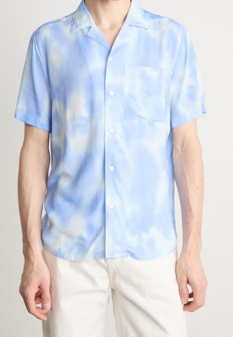 Kortärmad skjorta i ljusblått med tie-dye-mönster, slät tyg, en bröstficka och knäppning fram.