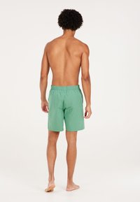 Groene zwembroek met een elastische tailleband, lichtgewicht stof en een ontspannen pasvorm, gezien vanaf de achterkant met een effen achtergrond.