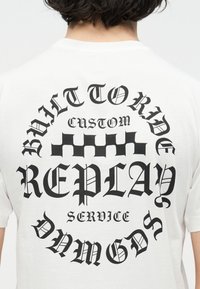 Άτομο που φοράει λευκό μπλουζάκι με μαύρο γοτθικό κείμενο και σχέδιο σαν σκακιέρα στο κέντρο της πλάτης.