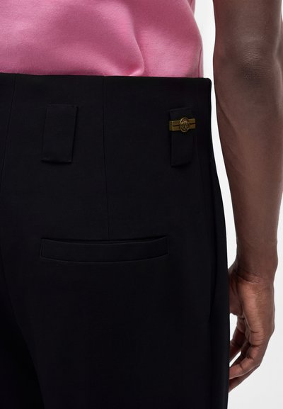 Gros plan d'une personne portant un pantalon noir avec un emblème doré à la taille et une chemise rose, montrant la poche arrière et une partie du bras.