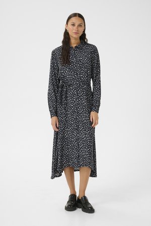 Robe à manches longues bleu marine avec un motif floral, tissu fluide et ceinture à nouer. Associée à des chaussures noires et à une boucles d'oreilles simples.