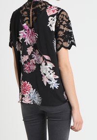 Blusa negra con estampado floral que presenta grandes flores coloridas, mangas cortas de encaje y un detalle de abertura en la parte de atrás. Tejido suave con una textura lisa.