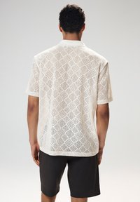 Fehér, rövid ujjú, csipkemintás ing gallérral, gyémántvágásos dizájnnal. Sötét rövidnadrággal párosítva.